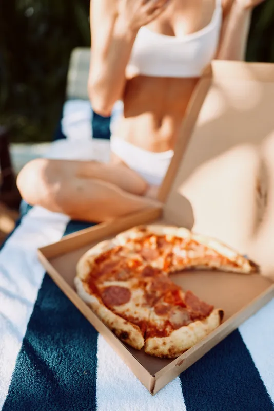 A sunny outdoor scene shows a relaxed person in a bikini top sitting on a blanket with an open pizza box, capturing a casual beachside meal, warm sunlight, and a carefree lifestyle moment.
