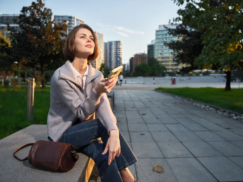 A cheerful professional woman sits on a bench in an urban park, looking at her phone with a smile, enjoying a calm, modern outdoor atmosphere.