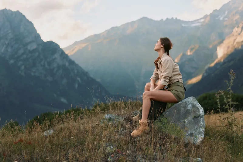Woman sitting on a rock in a mountain landscape, enjoying fresh air and quiet solitude. Casual outdoor style expresses adventure, wellness, and nature connection.