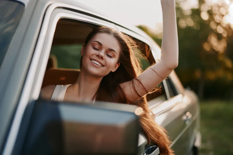 Smiling young woman enjoying a road trip with her hair flowing in the wind, captured during sunset, showcasing freedom and joy.