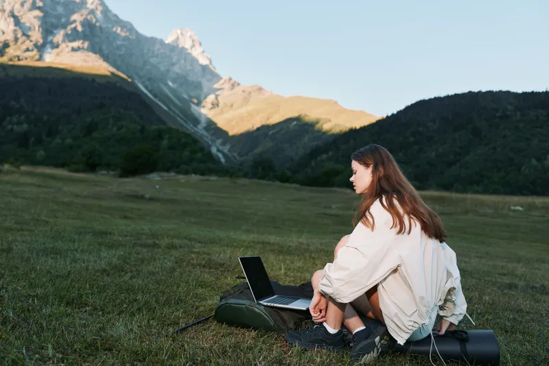 A lone traveler sits on a grassy field with a laptop, gazing at the screen as distant mountains rise, creating a calm, productive outdoor work atmosphere.