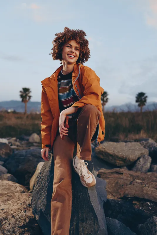 Woman lifestyle portrait, curly hair, smiling while walking outdoors in an orange jacket and casual denim striped shirt, standing on rocks, palms in background, cloudy sky, daylight