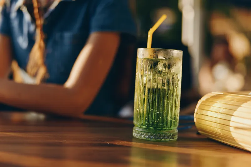 Refreshing green cocktail with ice and a straw on a wooden bar counter, warm ambient light and casual background with a blurred person in summer setting.