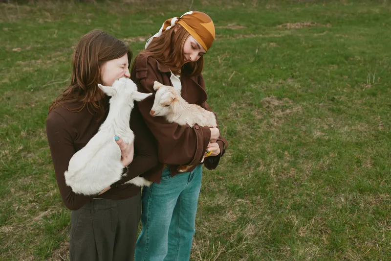 Two young women hold baby goats in their arms while standing on a green grassy field enjoying a peaceful lifestyle moment together outdoors.