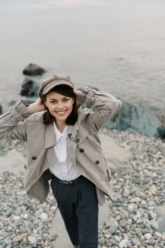 Smiling young woman wearing a hat and beige jacket stands on a rocky beach near calm water. Casual outfit and natural light create a relaxed and cheerful mood by the sea.