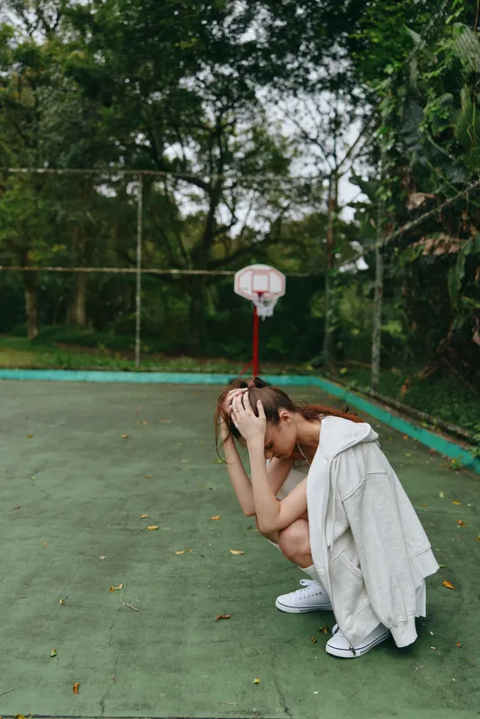 Young woman sitting on a basketball court, expressing sadness while wearing a casual outfit, surrounded by greenery and a basketball hoop in the background