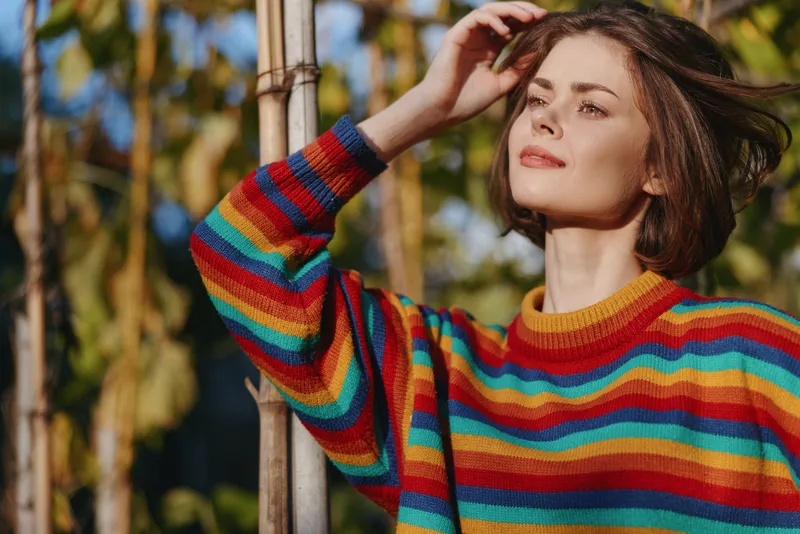 Woman young woman in a rainbow striped sweater outdoors in warm sunlight, smiling and touching short hair by bamboo; casual outfit, relaxed travel lifestyle portrait in nature.