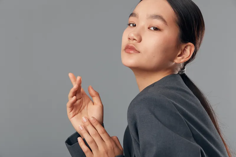 Young woman with long black hair tied back wearing dark gray jacket posing on gray background in studio with calm expression and elegant hand gesture in closeup portrait photography.