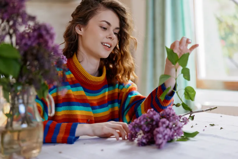 Cheerful woman indoors, arranging flowers on a bright table, colorful striped sweater, soft natural light, home hobby moment, relaxed mood, lush blooms in focus