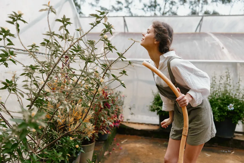 Young woman outdoors promoting sustainable gardening with a wooden hoe, wearing a white blouse and green overalls, surrounded by lush plants