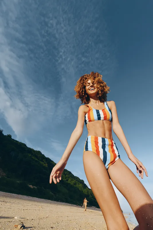 Woman beach in summer wearing swimsuit outdoors sunlight curly hair she walks with freedom, seaside vacation concept, warm sky over sand and green hills, low angle shot with sky today