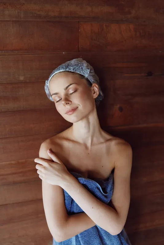 young woman wrapped in blue towel with shower cap poses peacefully against wooden wall, eyes closed and arms gently crossed, serene and calm atmosphere