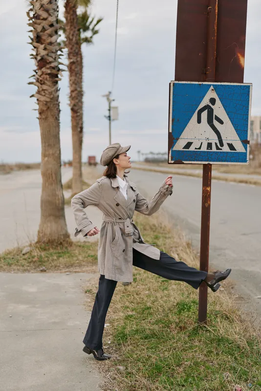 Young woman in casual autumn outfit poses near pedestrian crossing sign on roadside with palm trees in background. Concept of urban lifestyle, fashion, and outdoor activity.