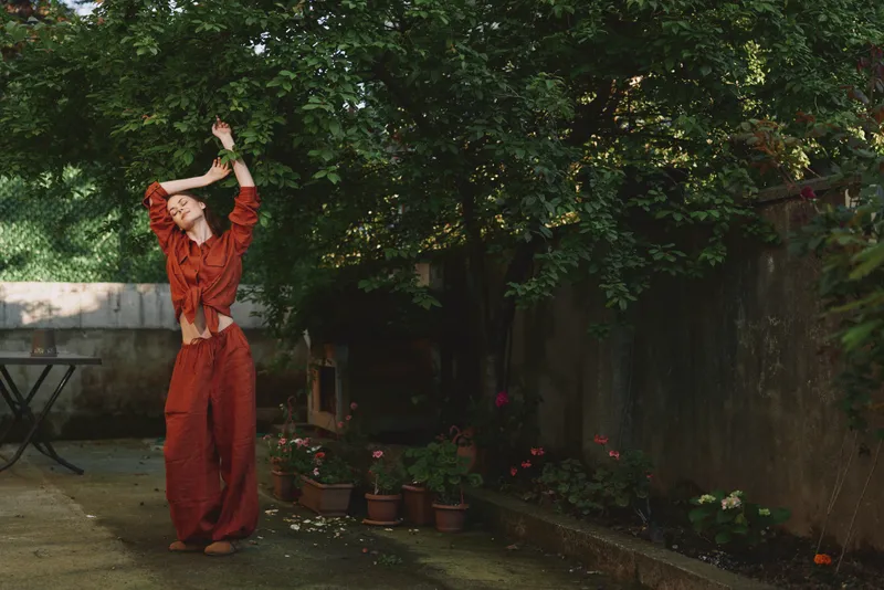 Woman in red outfit stretching arms outdoors in a sunny garden, a casual lifestyle scene that emphasizes wellness, confidence, creativity, and self care under warm natural light