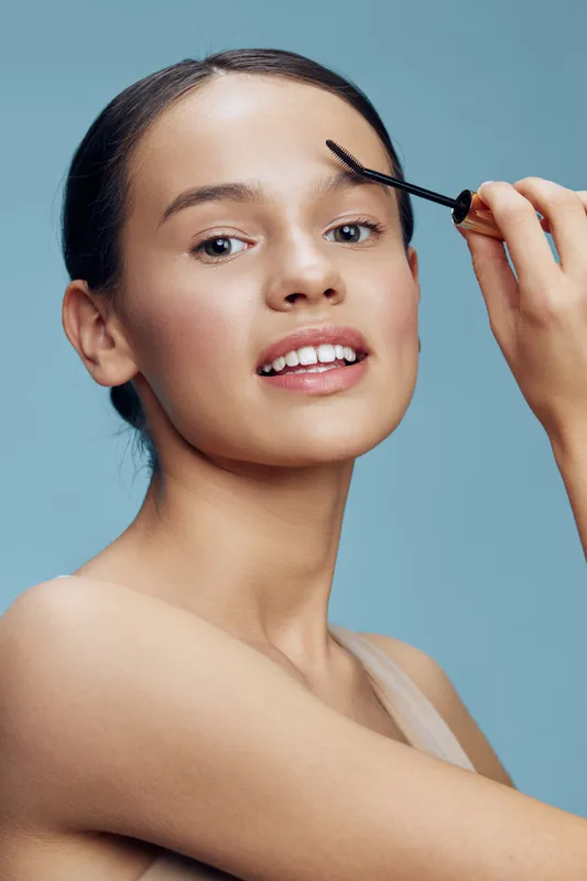 A confident young woman grooming her eyebrows with a brow gel, showcasing a natural beauty look against a serene light blue background
