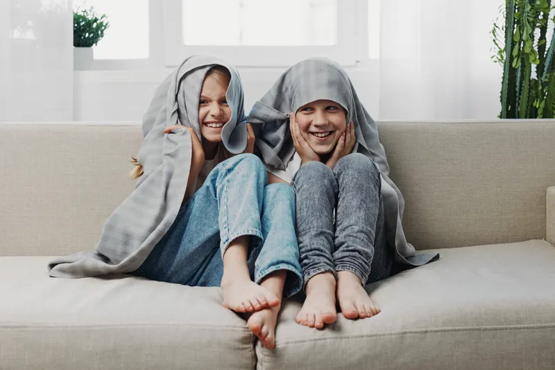Two happy children sitting on a sofa covered with a blanket, smiling and having fun indoors. Cozy home environment with natural light and casual clothes.