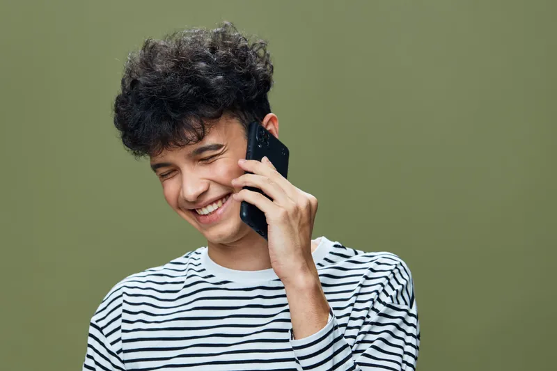 Smiling young man speaks on phone wearing striped shirt with curly hair, expressing happiness and relaxation against a plain olive green background.