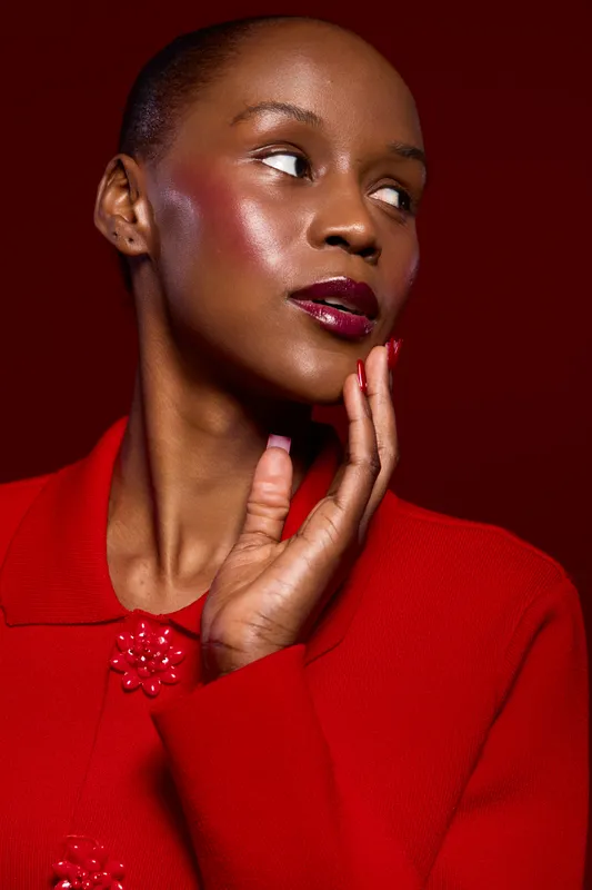 Portrait of a dark skinned African woman with glossy makeup wearing a red coat. She poses with hand near face on red background. Concept of beauty, fashion, elegance, and confidence.