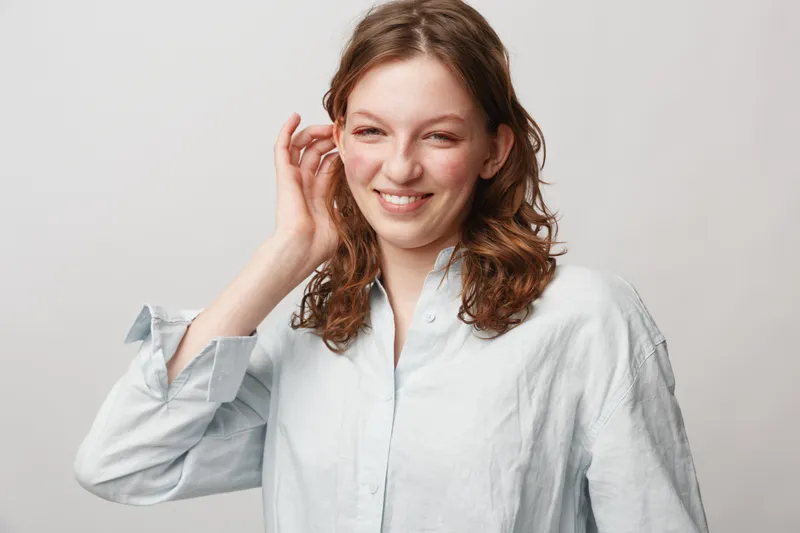 Smiling young woman with wavy hair wearing a light shirt touches her ear, looking at camera. Studio portrait on white background with natural expression and casual style.