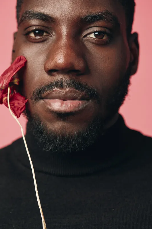 African man with dark skin and beard wearing black turtleneck holding red flower near face in creative conceptual visual style on pink background closeup portrait.