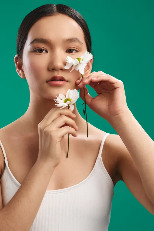 asian woman holds white flowers near her face in a clean studio setup with a vivid green backdrop, radiating calm elegance and natural beauty.