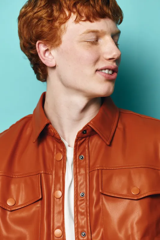 Young man with red hair wearing orange leather jacket and white shirt posing with closed eyes on blue background, fashion portrait and casual style for lifestyle.