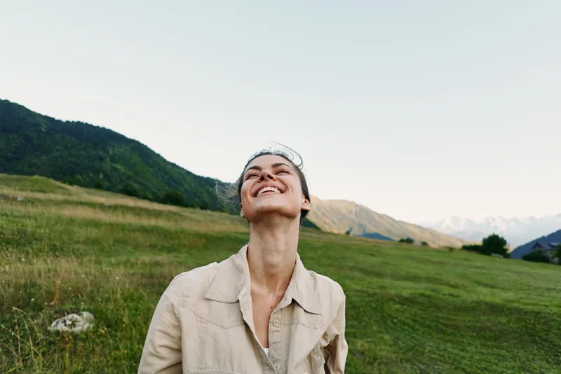 Cheerful woman outdoors in a beige jacket, smiling upward in a sunlit meadow with distant mountains, soft breeze, and vibrant greenery surrounding.