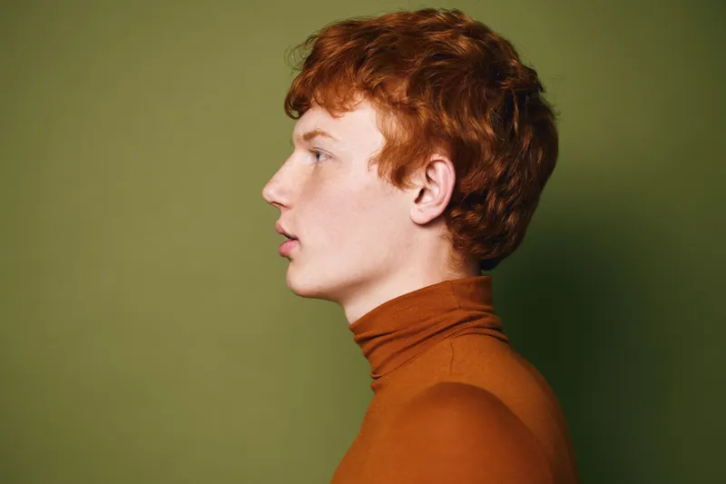 Profile portrait of young man with red hair wearing orange turtleneck against green background. Side view of serious male model with fair skin and short curly hairstyle in studio.