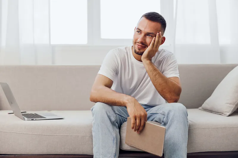 Thoughtful man sitting on a sofa, casually dressed in a white t-shirt and jeans, reflecting on ideas with a notebook and laptop nearby.