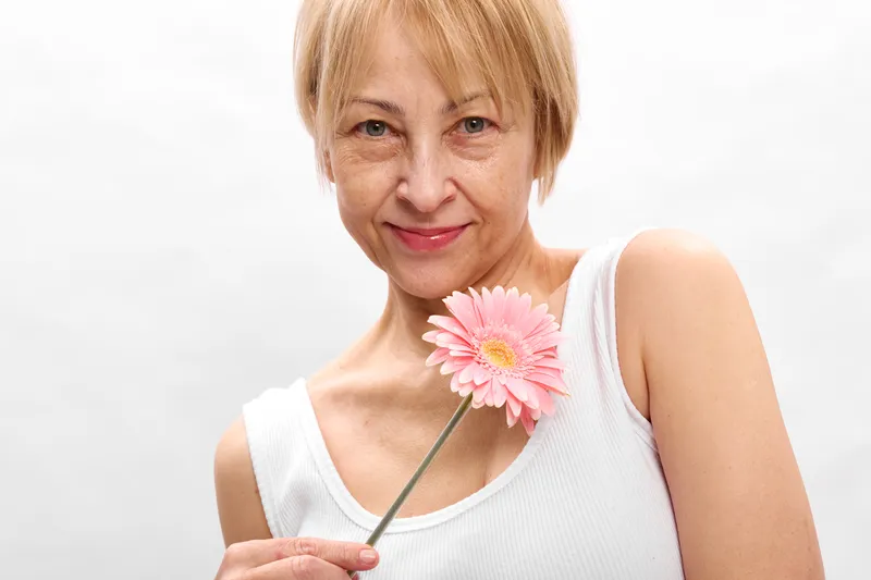 Woman elderly smiling flower pink spring portrait, white studio background, closeup of a relaxed lady holding gerbera blossom, soft light shows calm beauty.