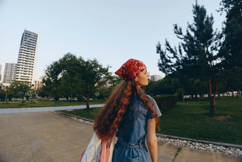 Lifestyle woman in modern boho-western denim street style with long hair and red bandana, captured in film color, walking outdoors near trees and urban buildings.