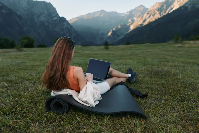 A woman sits on a rolled mat in a grassy field beside majestic mountains, working on a laptop outdoors. She wears casual clothes, focused on her screen, and enjoys quiet nature with modern technology.