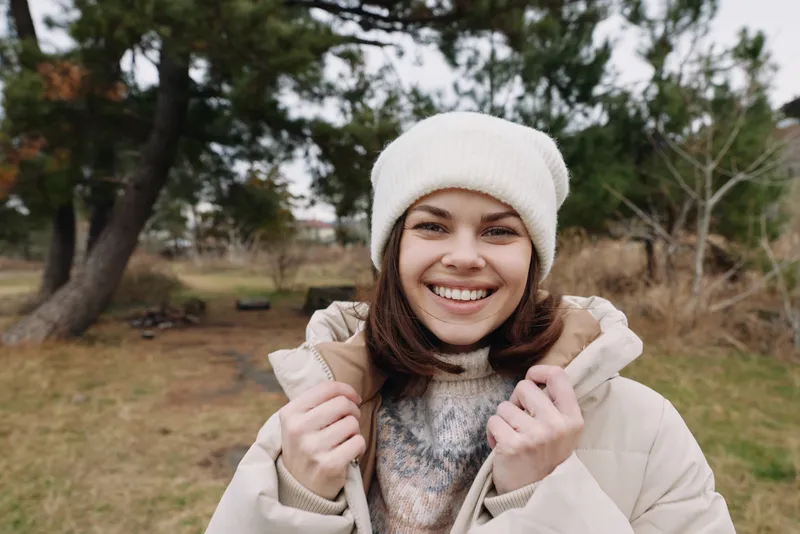 Woman in a warm coat and beanie smiles outdoors in a park with trees, creating a casual lifestyle portrait in chilly weather.
