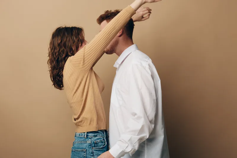 Couple in casual clothes embraces and kisses against beige background. Woman in sweater and jeans, man in white shirt. Concept of love, romance, intimacy, and relationship.