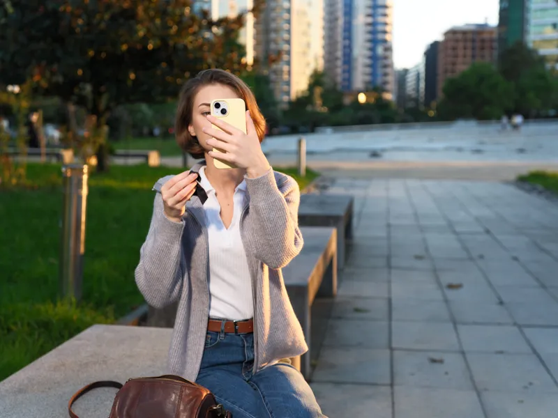 A cheerful woman takes a selfie in an outdoor urban park setting. She smiles warmly, wearing casual attire and a cardigan, creating a bright, confident moment amid modern city surroundings.