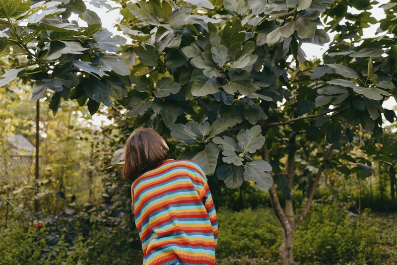 Woman in a colorful striped sweater leaning toward a tree in a suburban garden, young woman mid 20s with shoulder length hair in a casual outfit, peaceful lifestyle portrait outdoors.