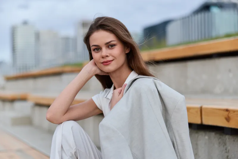 Young woman in casual light outfit smiling outdoors with modern urban background and natural soft light, warm autumn tones, relaxed confident pose
