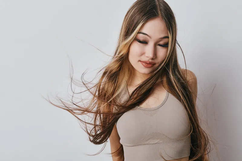 Young woman with long flowing hair wearing sleeveless top looking down against light background. Beauty, fashion, and natural style with soft expression and movement in hair.