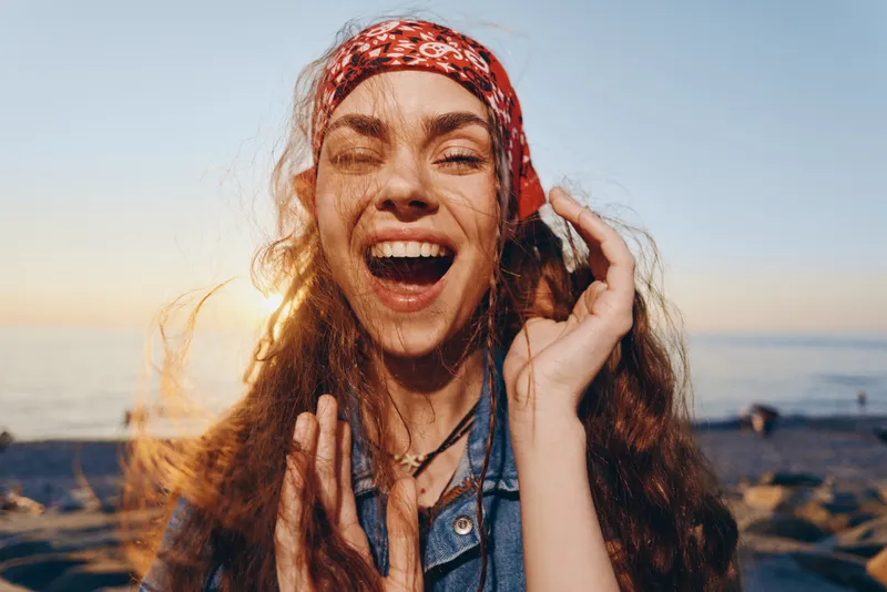 joyful woman laughing with curly hair and red headband outdoors at sunset, natural light, carefree expression, summer beach setting, warm tones