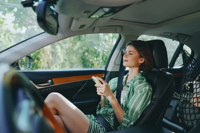 A young woman sits in car interior, looking ahead while using a smartphone. Natural daylight highlights her striped green dress and relaxed mood inside a modern cabin.