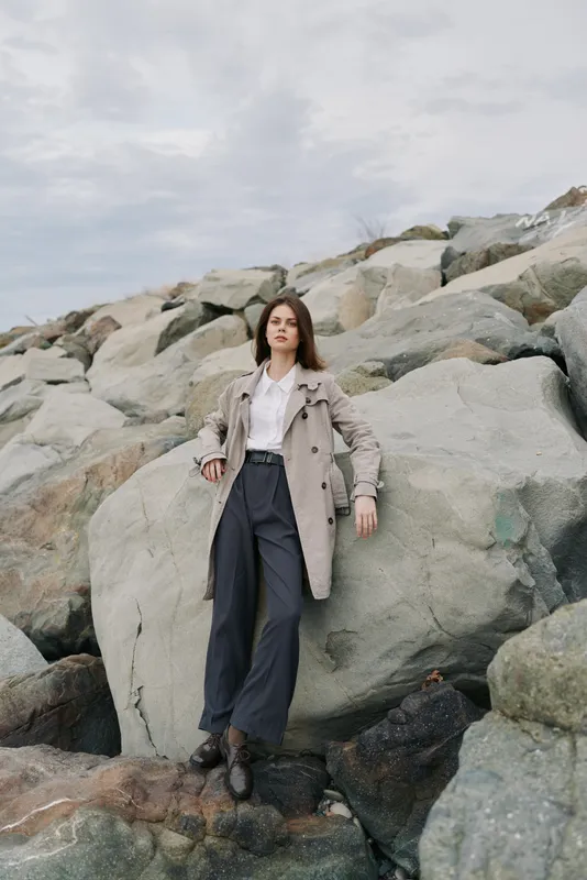 Young woman in casual outfit stands confidently on rocky shore with cloudy sky. Concept of fashion, nature, outdoor lifestyle, and serene mood by the sea.