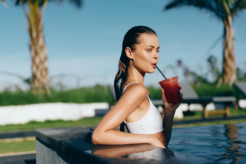 Young woman with long brunette hair drinks a red smoothie by the pool on a sunny day, enjoying a relaxed lifestyle moment with clear blue sky and palm trees in the background.