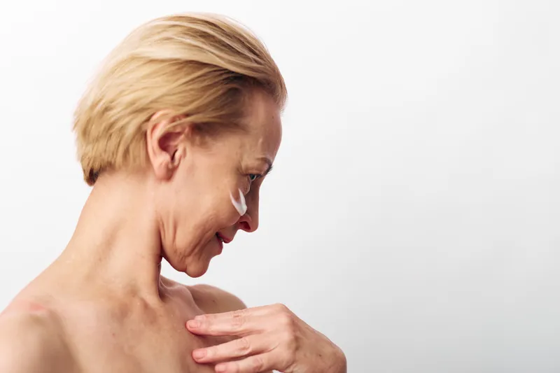 Mature woman with short blonde hair applies white cream on face and shoulder, showing skincare routine and self-care with soft natural lighting on white background.