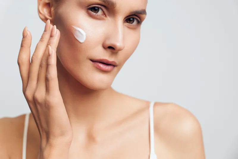 Young woman with clear skin applies white moisturizing cream on her cheek with fingers isolated on light background natural beauty skincare routine concept.