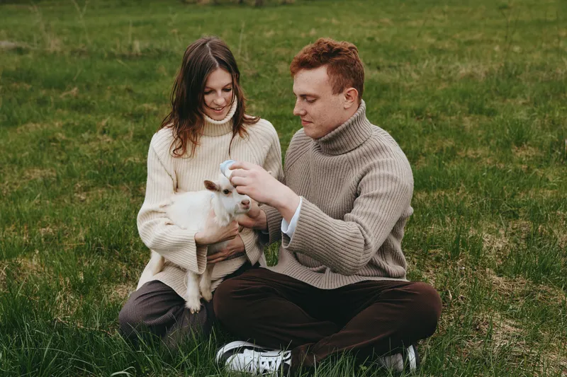 Couple sitting on grass, softly stroking a small goat kid. Caring moment outdoors, cozy sweater vibe, relaxed lifestyle. Tender connection, gentle touch, warm lighting, natural backdrop, peaceful