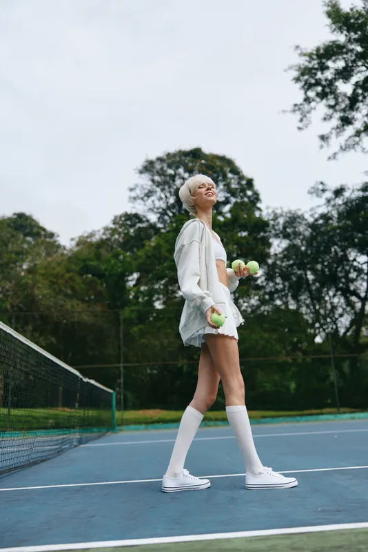 Young female athlete with short hair smiling on a tennis court, wearing a sporty white outfit, holding tennis balls, enjoying a sunny day outdoors