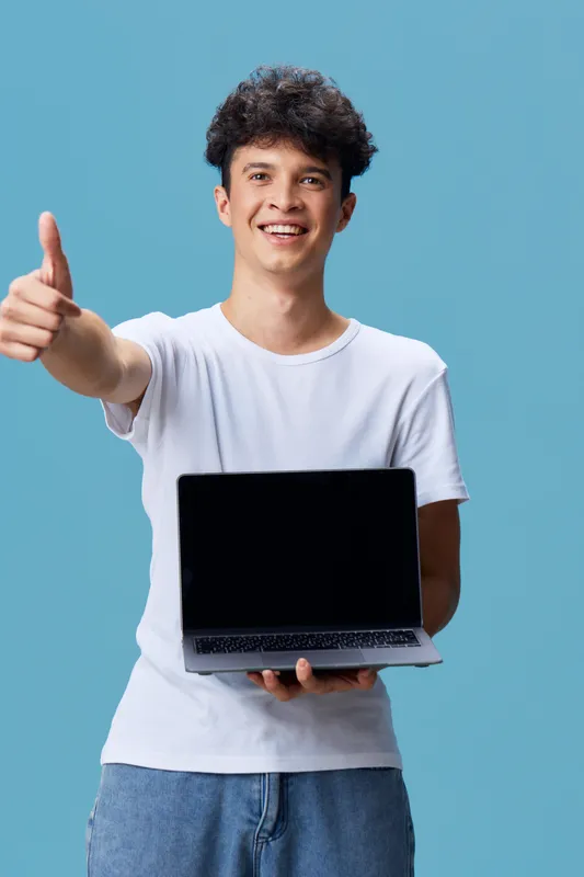 Smiling man holding laptop and giving thumbs up, wearing casual white t-shirt, standing against blue background with curly hair, cheerful and positive mood.