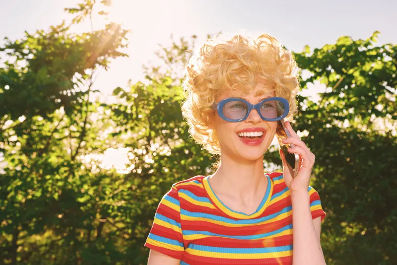 A blonde woman with curly hair in a striped colorful shirt smiles in a sunny outdoor setting, catching a retro lifestyle and carefree mood, framed by lush trees and bright light.