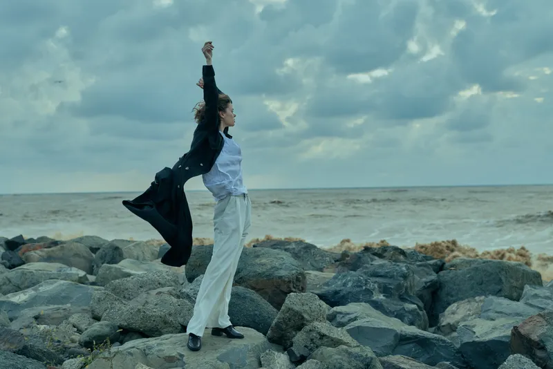 Woman on rocky shore with ocean horizon, wind in motion, celebrating achievement and freedom in outdoor attire, confident stance by water and sky and light.