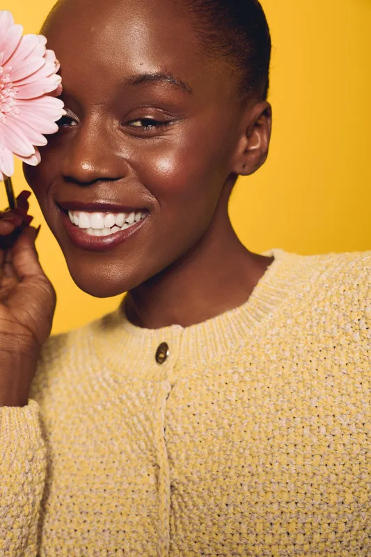 Smiling black woman with dark skin holds pink flower near face, wearing yellow sweater. Close-up portrait on yellow background. Concept of beauty, happiness, and natural skincare.